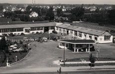 Schwarz-Weiß-Luftaufnahme eines Betriebsgeländes aus den 1950er Jahren. Zu sehen sind mehrere flache Gebäude mit großen Fenstern, zahlreiche geparkte Autos und eine Tankstelle im Vordergrund. Im Hintergrund erstreckt sich eine Wohnsiedlung mit Einfamilienhäusern.