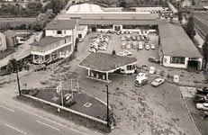 Schwarz-Weiß-Luftaufnahme eines Firmengeländes aus den 1960er Jahren. Das Gelände umfasst mehrere Gebäude, eine Tankstelle mit markantem Dach in der Mitte sowie zahlreiche geparkte Autos. Eine breite Straße verläuft vor dem Betriebsgelände, während im Hintergrund Wohnhäuser und Grünflächen zu sehen sind.