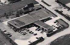 Schwarz-Weiß-Luftaufnahme eines Firmengeländes aus dem Jahr 1966. Das Gebäude hat eine rechteckige Struktur mit mehreren Parkplätzen und geparkten Autos darum herum. Das Gelände ist von Straßen umgeben, und im Hintergrund sind vereinzelte Häuser sowie offene Flächen sichtbar.