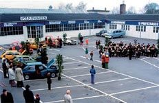 Farbige Aufnahme eines Firmengeländes aus dem Jahr 1975 während eines Polo-Turniers. Mehrere Autos sind auf einem Parkplatz ausgestellt, umgeben von dekorativen Pflanzen. Eine Menschenmenge, darunter Musiker mit Instrumenten, hat sich versammelt. Das Gebäude im Hintergrund trägt Beschriftungen wie 'Volkswagen' und 'Polo Klasse'.