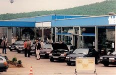 Aufnahme eines Autohauses aus den 1980er Jahren in Bad Driburg. Mehrere Audi-Fahrzeuge stehen vor dem modernen, mit Glasfassaden versehenen Gebäude, einige mit geöffneten Motorhauben. Menschen sind auf dem Gelände unterwegs, vermutlich bei einer Autoausstellung oder Verkaufsveranstaltung.