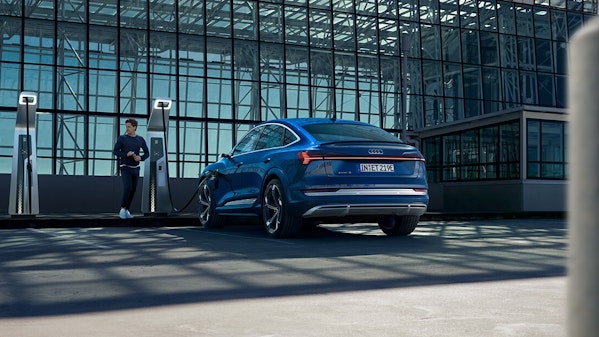 Blauer Audi e-tron Sportback lädt an einer modernen Ladestation vor einer Glasfassade, während ein Mann daneben steht – Elektromobilität in urbaner Architektur.