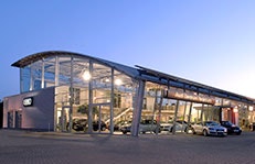 Moderne Audi-Autohalle bei Dämmerung im Jahr 2013. Das Gebäude hat eine markante Glasfassade mit geschwungenem Dach. Im Inneren sind mehrere Fahrzeuge ausgestellt, während draußen weitere Audis geparkt sind.