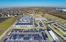 Luftaufnahme eines großen Autohauses in Rheda-Wiedenbrück aus dem Jahr 2017. Auf dem Dach des Hauptgebäudes sind zahlreiche Solarpaneele installiert. Der weitläufige Parkplatz ist mit vielen Fahrzeugen gefüllt, während im Hintergrund eine ländliche Umgebung mit Wohnhäusern zu sehen ist.