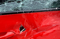Nahaufnahme eines stark beschädigten roten Autos mit zerbrochener Windschutzscheibe und einer großen Beule in der Karosserie. Die Schäden wurden vermutlich durch einen Tornado verursacht.